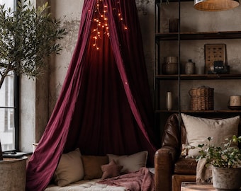 Könnte beinhalten: Ein tiefburgunderroter Stoffhimmel, der von der Decke hängt, mit einer konischen Spitze und fließenden, bodenlangen Vorhängen. Im Inneren des Baldachins hängt eine Lichterkette mit warmweißem Licht über einem beigen Bodenkissen mit Kissen.