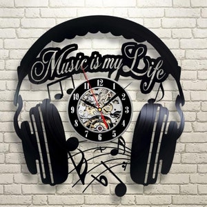 Puede incluir: Reloj de vinilo negro con diseño de auriculares y el texto "Music is my Life". La esfera del reloj es un círculo plateado con números negros y un mecanismo de relojería visible.