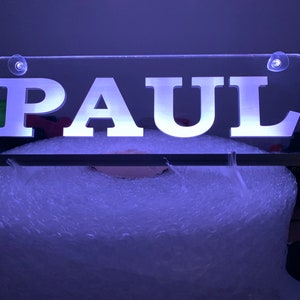 Puede incluir: Un letrero de acrílico transparente con el nombre "PAUL" en letras blancas. El letrero está iluminado desde atrás con una luz blanca y tiene dos pequeñas ventosas transparentes en la parte superior.