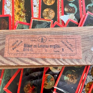 May include: A collection of vintage magic lantern slides with red borders and circular illustrations. The slides are arranged in a wooden box, with a label that reads "Bilder zu Laterna magica." The images depict various scenes and characters.