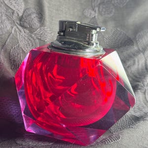 May include: A red, faceted lighter with a chrome top. The body of the lighter is a translucent, geometric shape with a swirling pattern inside. The lighter is set against a dark, textured background.