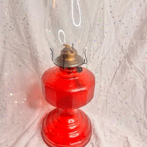 May include: A red glass oil lamp with a clear glass chimney. The lamp has a textured, faceted base and a brass-coloured burner assembly. The lamp is designed to hold oil and provide light.