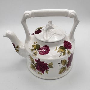 May include: White ceramic teapot with a bamboo-style handle and lid. The teapot is decorated with burgundy roses, white flowers, and green leaves. The spout and handle are white.
