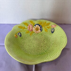 May include: A light green ceramic bowl with a textured, pebbled surface. The rim features a raised, moulded floral design with pink and white flowers, yellow leaves, and small blue buds. The bowl has a scalloped edge and a glossy finish.