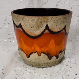 May include: A ceramic planter featuring a speckled beige base and rim with a wide, vibrant orange band around the centre. The orange section is defined by dark brown, jagged, drip-like outlines that create a distinct contrast against the lighter tones.