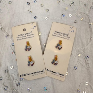 May include: Two sets of Peter Rabbit The Rabbit Postman buttons from the Beatrix Potter Collection by JHB International. Each set includes two small, colorful rabbit-shaped buttons attached to a rectangular beige card with printed text.