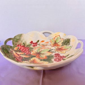 Peut inclure: Un bol de service en céramique couleur crème avec deux anses intégrées et un bord festonné. L'intérieur présente un motif peint de baies rouges et de feuilles aux couleurs automnales dans des tons orange, jaune et vert, avec un motif en relief de type treillis sur les bords.