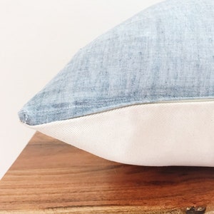 May include: A light blue pillow with a white border. The pillow is made of a soft, textured fabric and is sitting on a wooden surface.