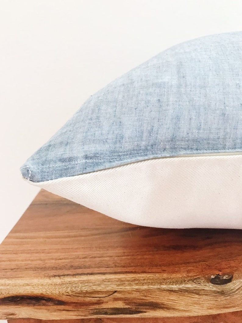 May include: A light blue pillow with a white border. The pillow is made of a soft, textured fabric and is sitting on a wooden surface.