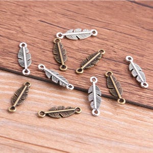 May include: A selection of metal feather charms in silver and bronze hues. Each charm has a detailed feather design with a loop at each end for attaching to jewellery or crafts. The charms are scattered on a wooden surface.