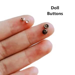 Puede incluir: Primer plano de una mano sosteniendo tres pequeños botones de muñeca. Dos son blancos y uno es marrón.