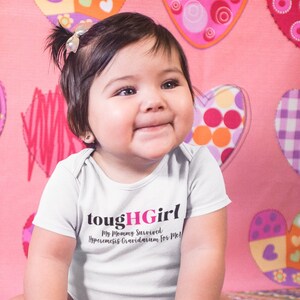 Puede incluir: Un bebé con un body blanco con el texto "toughHGirl My Mommy Survived Hyperemesis Gravidarum for Me!" El bebé está sentado en una manta rosa y blanca con estampado de corazones y juguetes coloridos.