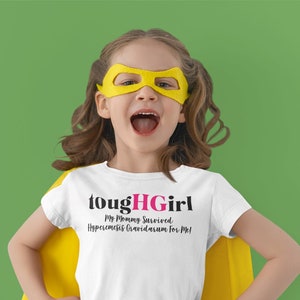 Puede incluir: Una niña pequeña con una camiseta blanca con el texto "toughHGirl My Mommy Survived Hyperemesis Gravidarum for Me!" Lleva una capa amarilla y una máscara amarilla.