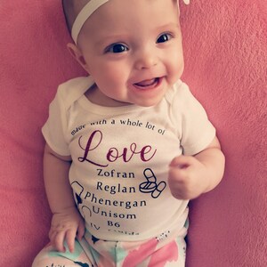 Könnte beinhalten: Ein weißer Strampler mit dem Text "Made with a whole lot of Love" und einer Liste von Medikamenten, darunter Zofran, Reglan, Phenergan, Unisom, B6 und IV Fluids. Ein Baby trägt den Strampler und lächelt.
