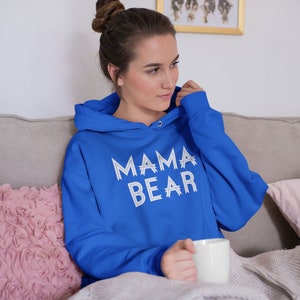 May include: A woman wearing a royal blue hoodie with the text "MAMA BEAR" in white on the front. She is sitting on a couch with a pink pillow and a white mug in her hand.
