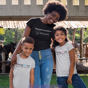 Könnte beinhalten: Eine dreiköpfige Familie trägt graue T-Shirts mit dem Text "toughGUY" und "toughGIRL" sowie den Worten "Hyperemesis Gravidarum Survivor". Die Familie steht in einem grasbewachsenen Bereich mit einem Holzzaun und einem braunen Pferd im Hintergrund.