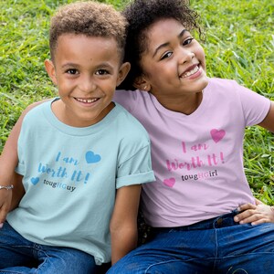 Könnte beinhalten: Zwei Kinder tragen blaue Jeans und T-Shirts mit dem Text "Ich bin es wert!" und den Worten "tough guy" und "tough girl" darauf gedruckt. Die Kinder sitzen auf einer grünen Grasfläche.