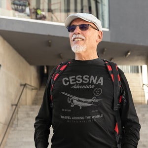 May include: Black long-sleeve shirt with a Cessna Adventure graphic. The shirt features a vintage airplane illustration and the text "Travel Around The World, Retro Airlines". The person is wearing gray cargo pants and a white cap.