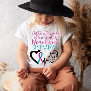 Puede incluir: Una camiseta blanca con una cinta rosa y azul, un corazón y un gráfico de latido del corazón. El texto en la camiseta dice "Difficult roads often lead to beautiful destinations".