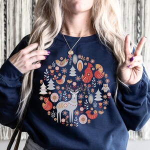 May include: A navy blue sweatshirt with a circular design featuring a deer, foxes, birds, and trees in a Scandinavian folk art style.