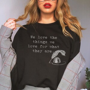 May include: A black t-shirt with the text "We love the things we love for what they are." in white lettering. The shirt features a small illustration of a quill pen and inkwell.