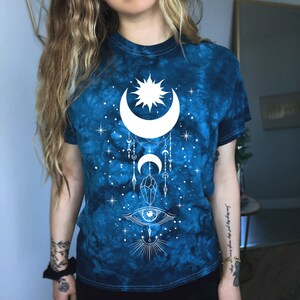 May include: A blue tie-dye t-shirt with a white moon, sun, and eye design. The design also includes stars and crystals.