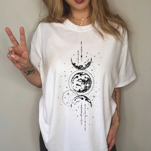 May include: White t-shirt featuring a black graphic of moon phases and celestial elements. The design includes crescent moons, full moons, stars, and linear details. The shirt has a crew neck and short sleeves.