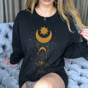May include: Black sweatshirt with a gold celestial design featuring a crescent moon, sun, stars, and a crystal. The design also includes an eye with a sunburst radiating from it.