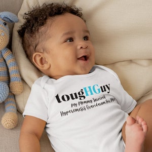 Puede incluir: Un bebé con un body blanco con el texto "toughGUY My Mommy Survived Hyperemesis Gravidarum for Me". El bebé está acostado en un sofá marrón claro con dos animales de peluche de crochet.