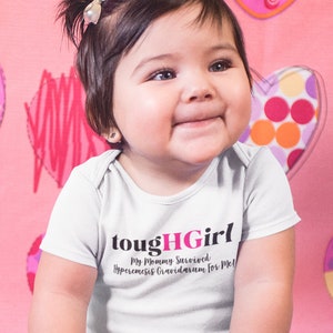 Könnte beinhalten: Ein Baby Mädchen trägt einen weißen Strampler mit dem Text "toughHGirl My Mommy Survived Premature Ovarian Failure for Me!"  Das Baby sitzt auf einer rosa-weißen Decke mit Herzmuster und Spielzeug um sich herum.