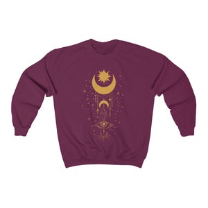 May include: Burgundy sweatshirt with a gold celestial design featuring a crescent moon, sun, stars, and an eye.