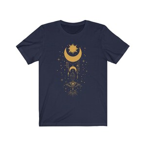 May include: A navy blue t-shirt with a gold crescent moon, sun, and eye design. The design features a crescent moon with a sun inside, a dangling eye, and stars.