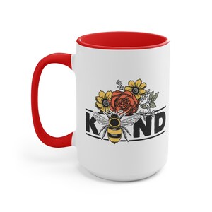 Op de afbeelding: Witte keramische mok met een rode rand en handvat. De mok heeft een zwart-witte afbeelding van een bij met bloemen en het woord "KIND".