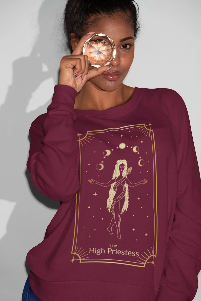 Op de afbeelding: Bordeauxkleurige trui met een gouden illustratie van de tarotkaart de Hogepriesteres. De kaart toont een vrouw met lang haar, een slang en een halve maan.