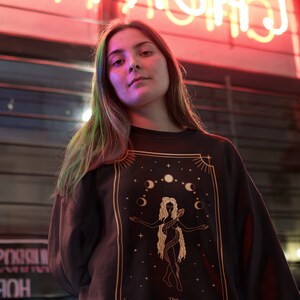 Op de afbeelding: Een vrouw draagt een zwart shirt met lange mouwen met een gouden afbeelding van een vrouw met een slang en manen. De afbeelding is omgeven door een gouden frame.