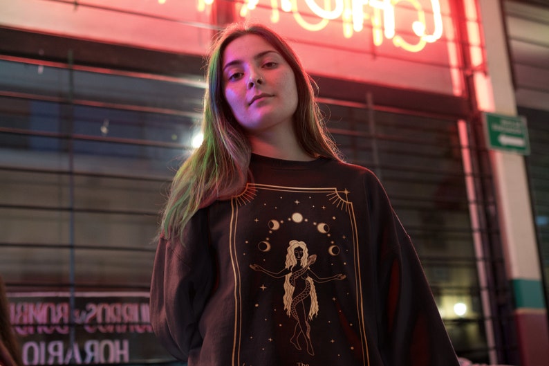 Op de afbeelding: Een vrouw draagt een zwart shirt met lange mouwen met een gouden afbeelding van een vrouw met een slang en manen. De afbeelding is omgeven door een gouden frame.