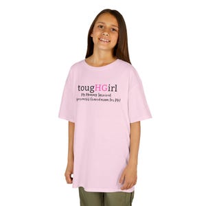 Pode incluir: Uma t-shirt rosa claro com o texto "toughHGIRL" em preto e rosa, com o texto "My Mommy Survived Hyperemesis Gravidarum For Me!" por baixo. A t-shirt é usada por uma pessoa jovem.
