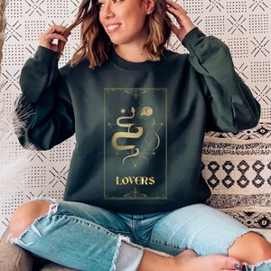 Puede incluir: Una sudadera verde oscuro con un gr&aacute;fico dorado de una serpiente y la palabra "LOVERS" en la parte delantera.