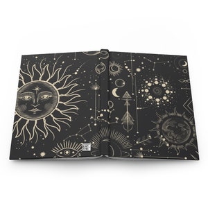 May include: An open book with a black cover and a white design of celestial bodies, including a sun, moon, stars, and constellations. The design is repeated on both pages of the book.