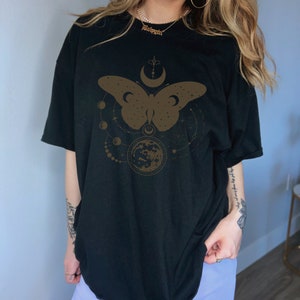 May include: Black oversized t-shirt with a brown butterfly graphic design. The butterfly is surrounded by a crescent moon, stars, and a circle with a crescent moon inside. The text "Beloved" is visible on a gold chain necklace.