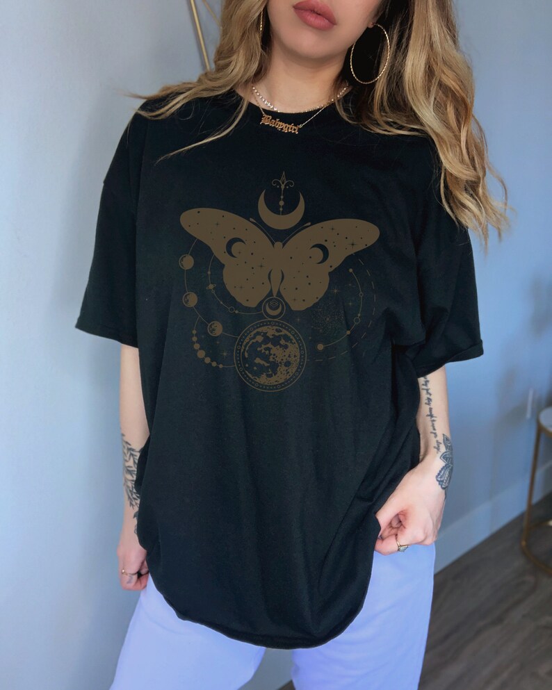 May include: Black oversized t-shirt with a brown butterfly graphic design. The butterfly is surrounded by a crescent moon, stars, and a circle with a crescent moon inside. The text "Beloved" is visible on a gold chain necklace.