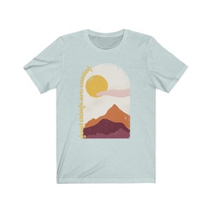 Puede incluir: Camiseta azul claro con un gr&aacute;fico de una cordillera, un sol y una mano que sale de una nube. El texto "good things are coming" est&aacute; escrito verticalmente en el lado izquierdo del gr&aacute;fico.