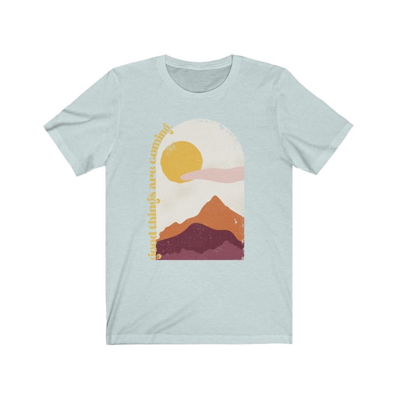 Puede incluir: Camiseta azul claro con un gr&aacute;fico de una cordillera, un sol y una mano que sale de una nube. El texto "good things are coming" est&aacute; escrito verticalmente en el lado izquierdo del gr&aacute;fico.