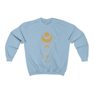 May include: A light blue sweatshirt with a gold celestial design featuring a crescent moon, a sun, and an eye. The design is surrounded by stars.