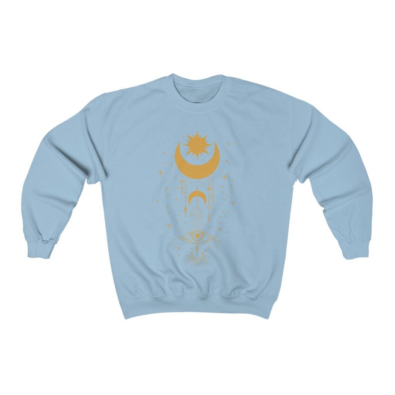 May include: A light blue sweatshirt with a gold celestial design featuring a crescent moon, a sun, and an eye. The design is surrounded by stars.