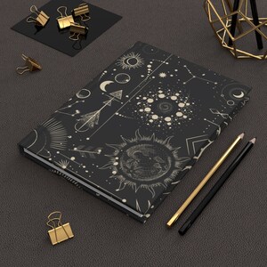 May include: A black notebook with a celestial design featuring moons, stars, and sun symbols in a cream color. The notebook is lying on a dark gray textured surface with gold paper clips and pencils.