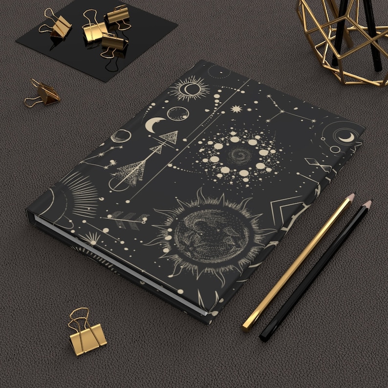 May include: A black notebook with a celestial design featuring moons, stars, and sun symbols in a cream color. The notebook is lying on a dark gray textured surface with gold paper clips and pencils.