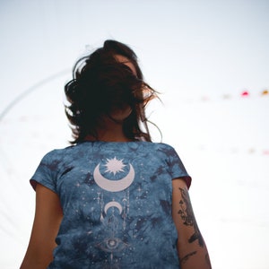 May include: A woman wearing a blue tie-dye t-shirt with a white moon, star, and eye design. The shirt has a faded, vintage look.