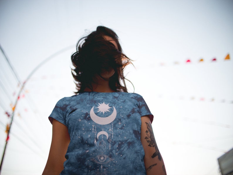 May include: A woman wearing a blue tie-dye t-shirt with a white moon, star, and eye design. The shirt has a faded, vintage look.