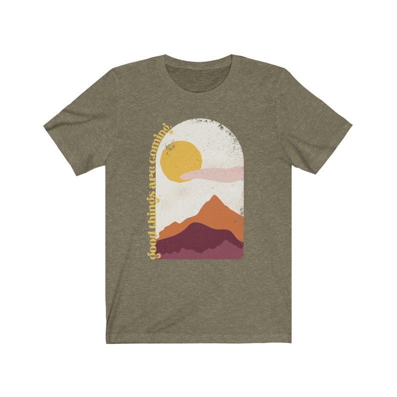 Puede incluir: Una camiseta verde con un gr&aacute;fico de una cordillera y un sol con el texto "good things are coming" en una fuente retro.